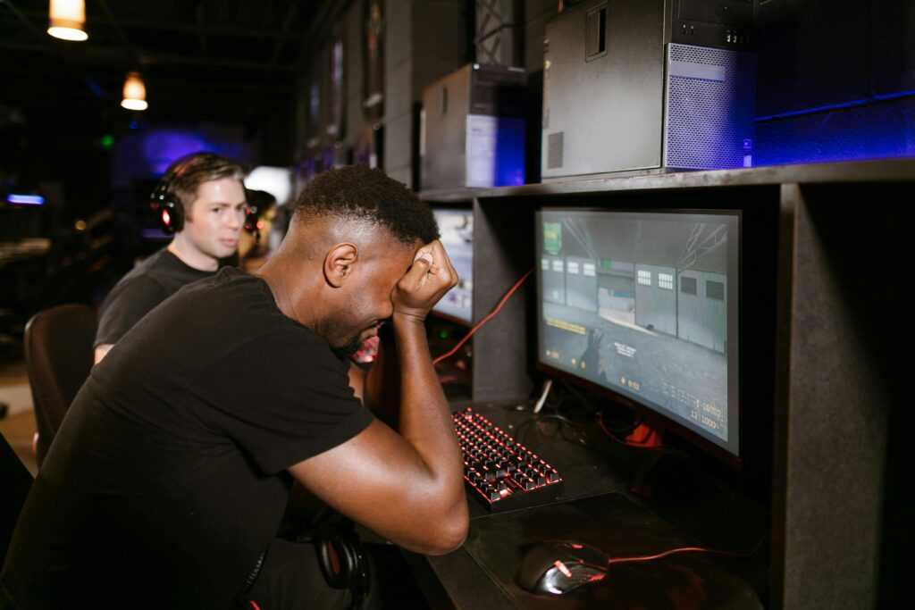 Two adult men experiencing disappointment while playing PC games in a modern indoor gaming arcade setting.
